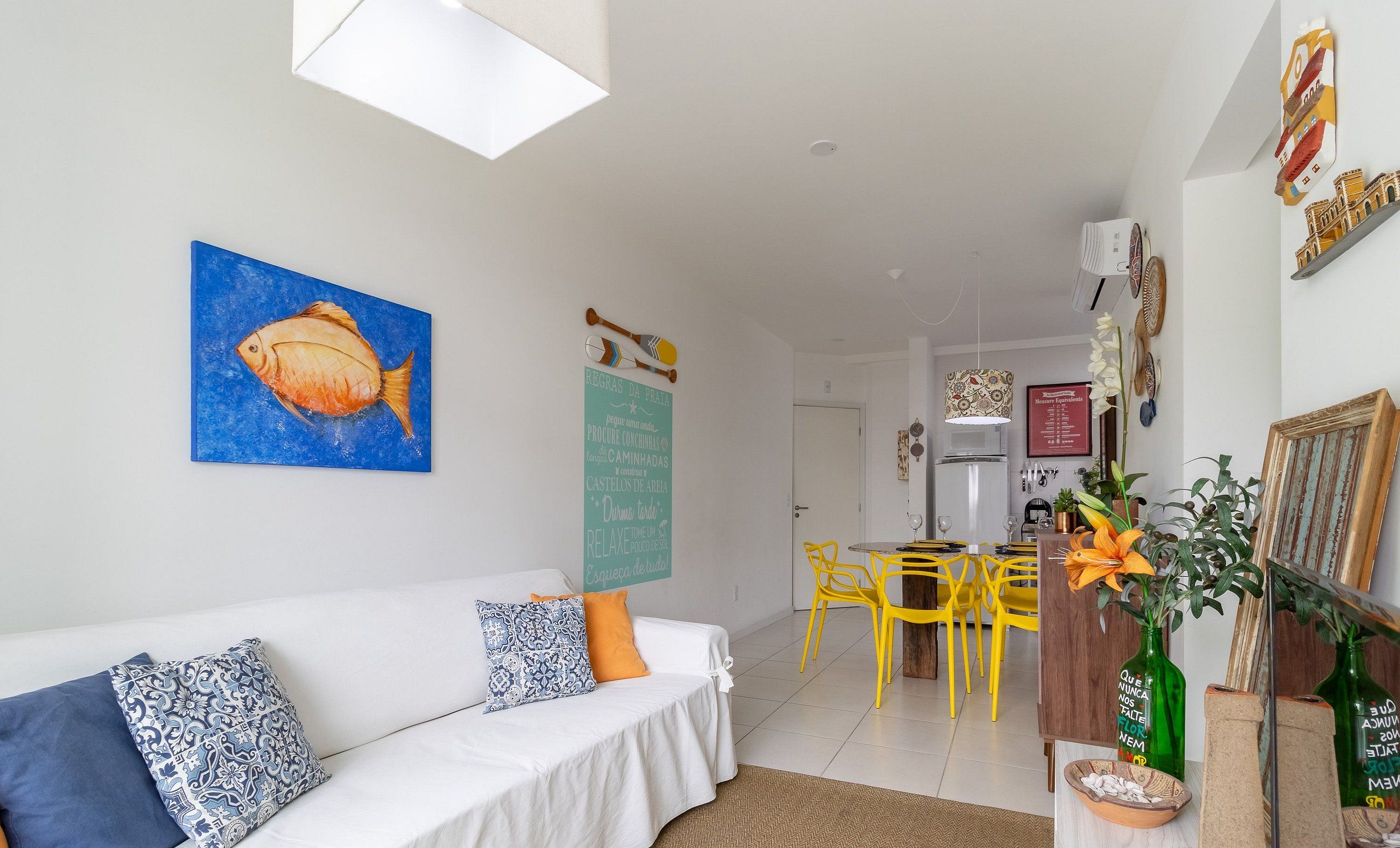 Un salon avec un canapé blanc, un tableau représentant des poissons bleus et un coin repas avec des chaises jaunes.