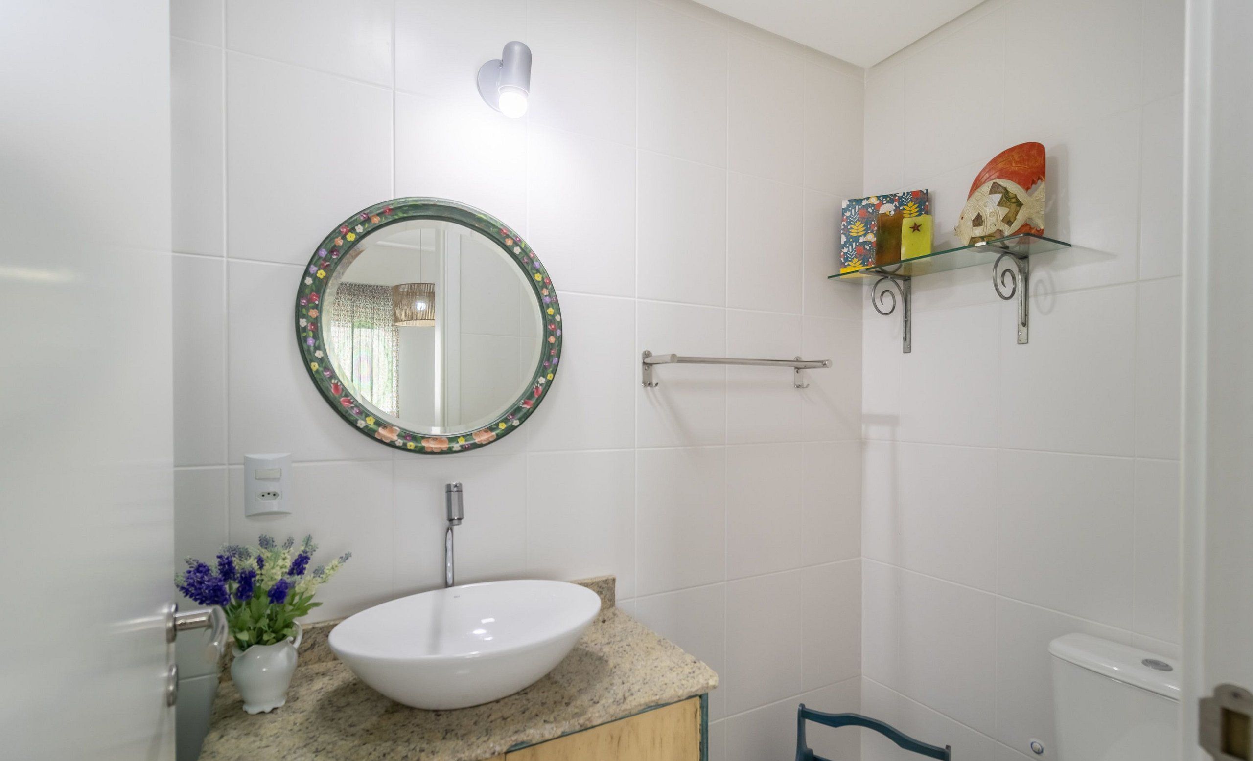 A bathroom with a round mirror, a white sink, a towel rack, and a shelf with decorative items.