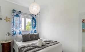 A bedroom with a bed, a bedside table, a lamp, a starfish decoration, and a window with patterned curtains. - Moderno Apto c/ Piscina em Canasvieiras RCP202 (Florianópolis)