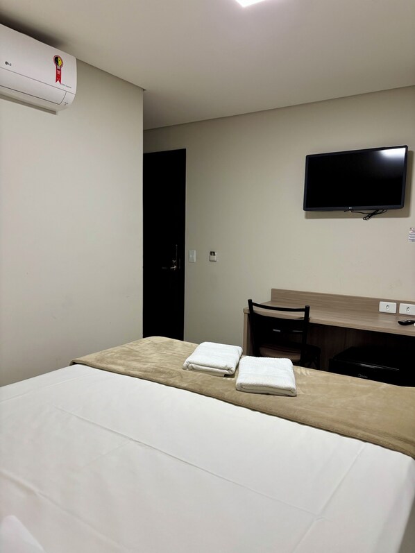 A hotel room with a bed, a desk, a chair, a flat-screen TV, and an air conditioner.