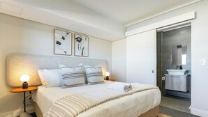 A neatly arranged hotel room with a bed, bedside lamps, a small bedside table, and a bathroom visible through an open door. - Century City Escape | Walk to Shops and Views (Cape Town)