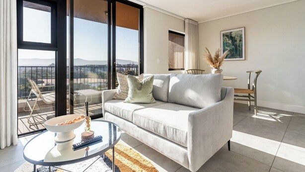 A modern living room with a sofa, a glass coffee table, and a view of the outdoors. - Century City Escape | Walk to Shops and Views (Cape Town)