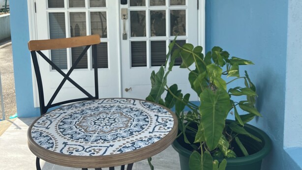A blue exterior wall with a white door marked with the number 2, a round table with a patterned tablecloth, a wooden chair, and a potted plant.