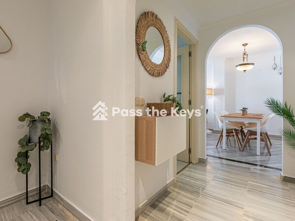 Un intérieur moderne avec un couloir menant à un espace repas. On y trouve un meuble en bois surmonté d’un miroir, une plante en pot et une table à manger avec des chaises.