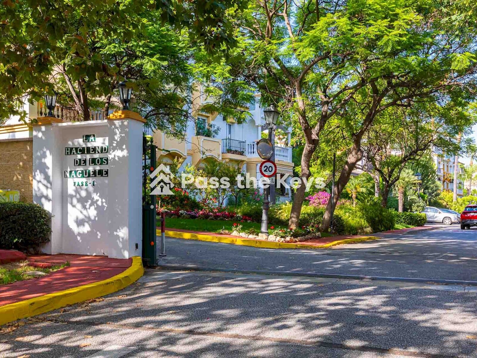 A street with a speed limit sign of 20, a gated entrance with a sign reading "LA HACIENDA DE LOS NAGUELES FASE I", and a building with balconies in the background.