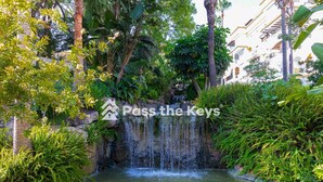 A lush garden with a waterfall and a pond, surrounded by tropical plants and a white building in the background.