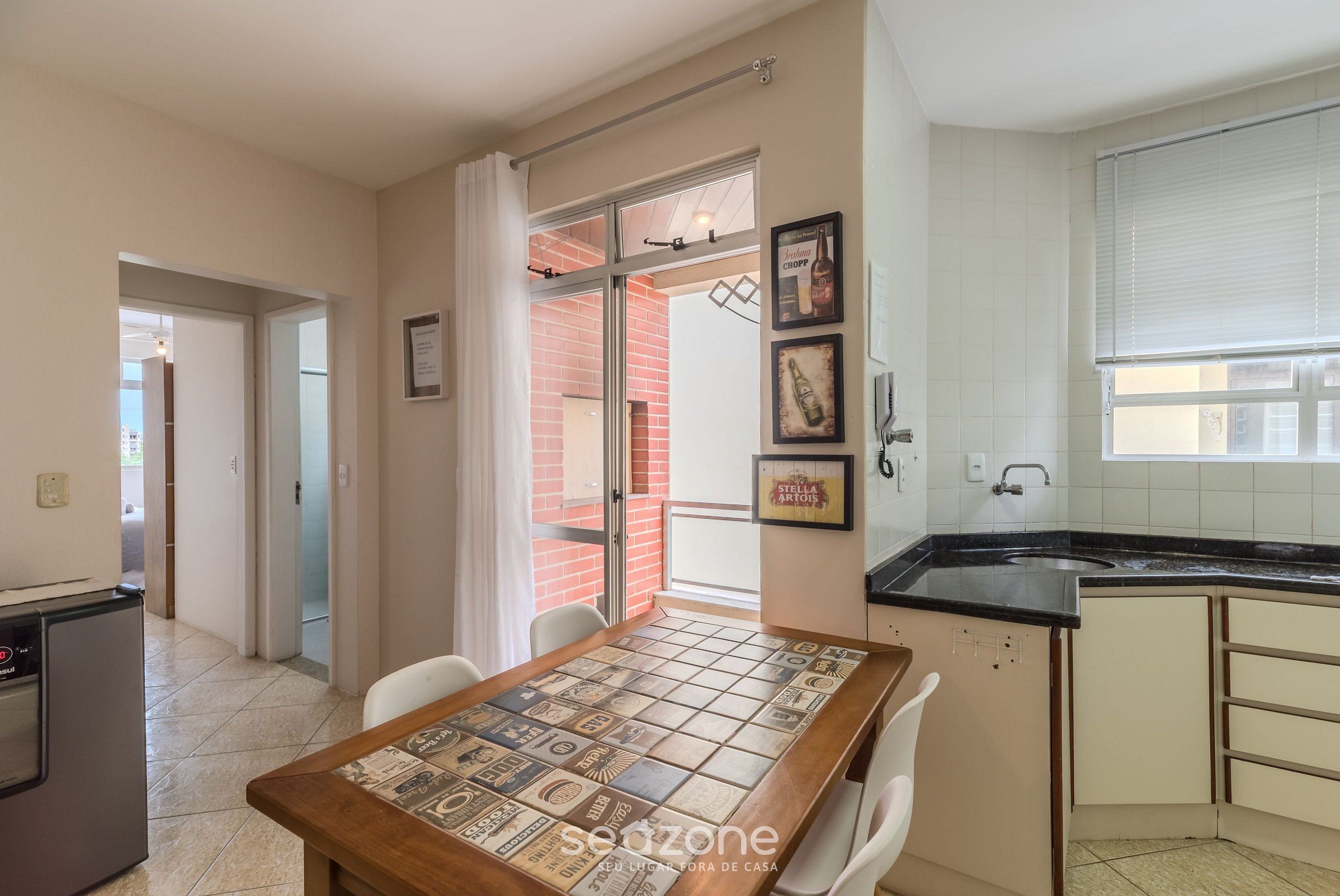 Une cuisine avec un plan de travail noir, un évier et une fenêtre avec des stores. On y trouve une table à manger avec des chaises et un mur recouvert de carreaux.