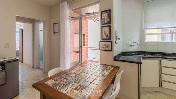 A kitchen with a black countertop, a sink, and a window with blinds. There is a dining table with chairs and a tiled backsplash.