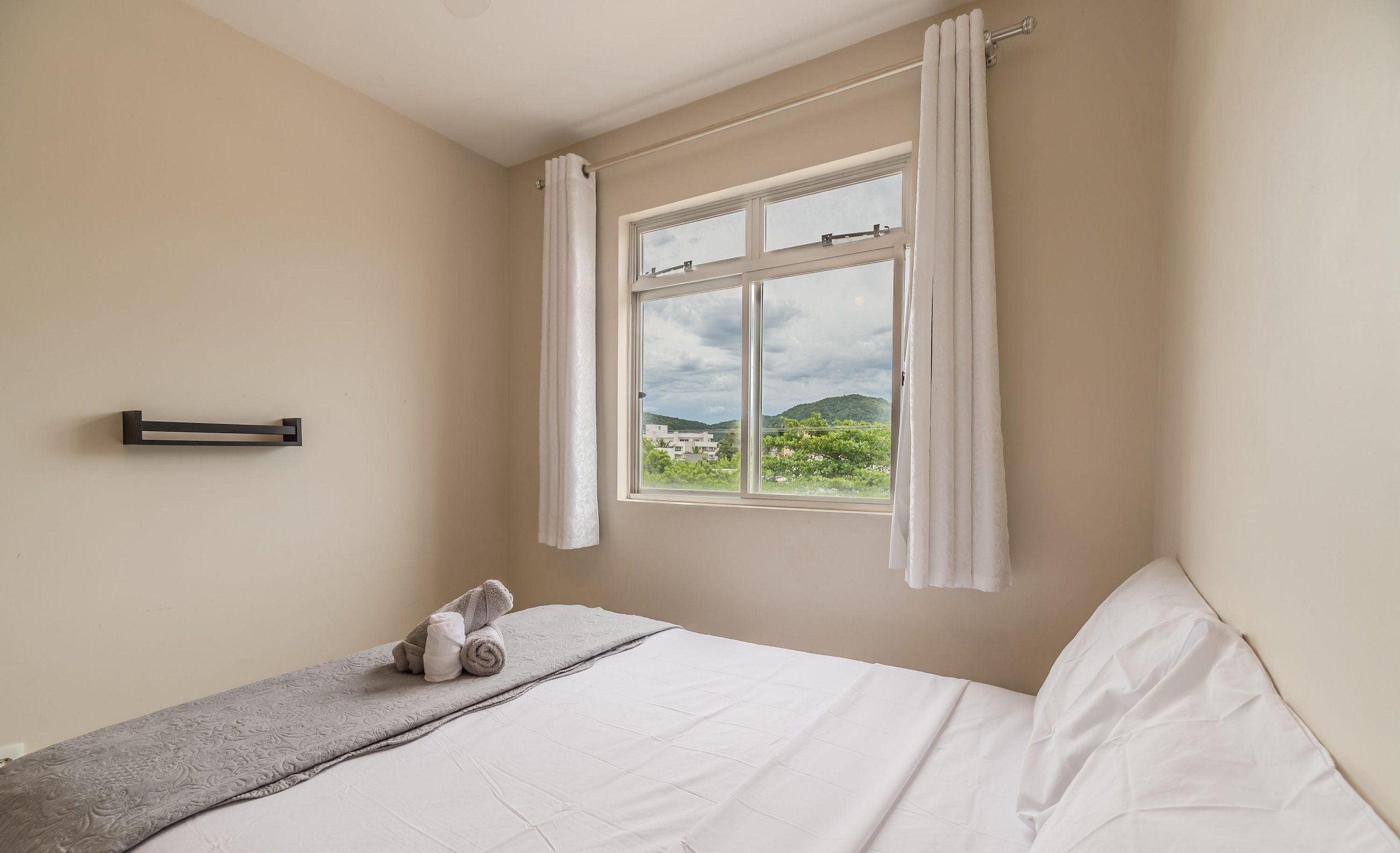 Un dormitorio con cama, ventana con cortinas, toalla sobre la cama y un toallero de pared.