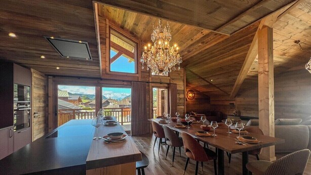 A spacious dining area with a large wooden table set for six, surrounded by chairs. A chandelier hangs from the ceiling, and the room features exposed wooden beams and a large window offering a view of the outside. - New Agora 202 Duplex | Central close to the slopes (Les Gets)