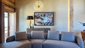 A cozy living room with a grey sectional sofa, a chandelier, a framed picture of a snowy landscape, and a wooden ceiling. - New Agora 202 Duplex | Central close to the slopes (Les Gets)