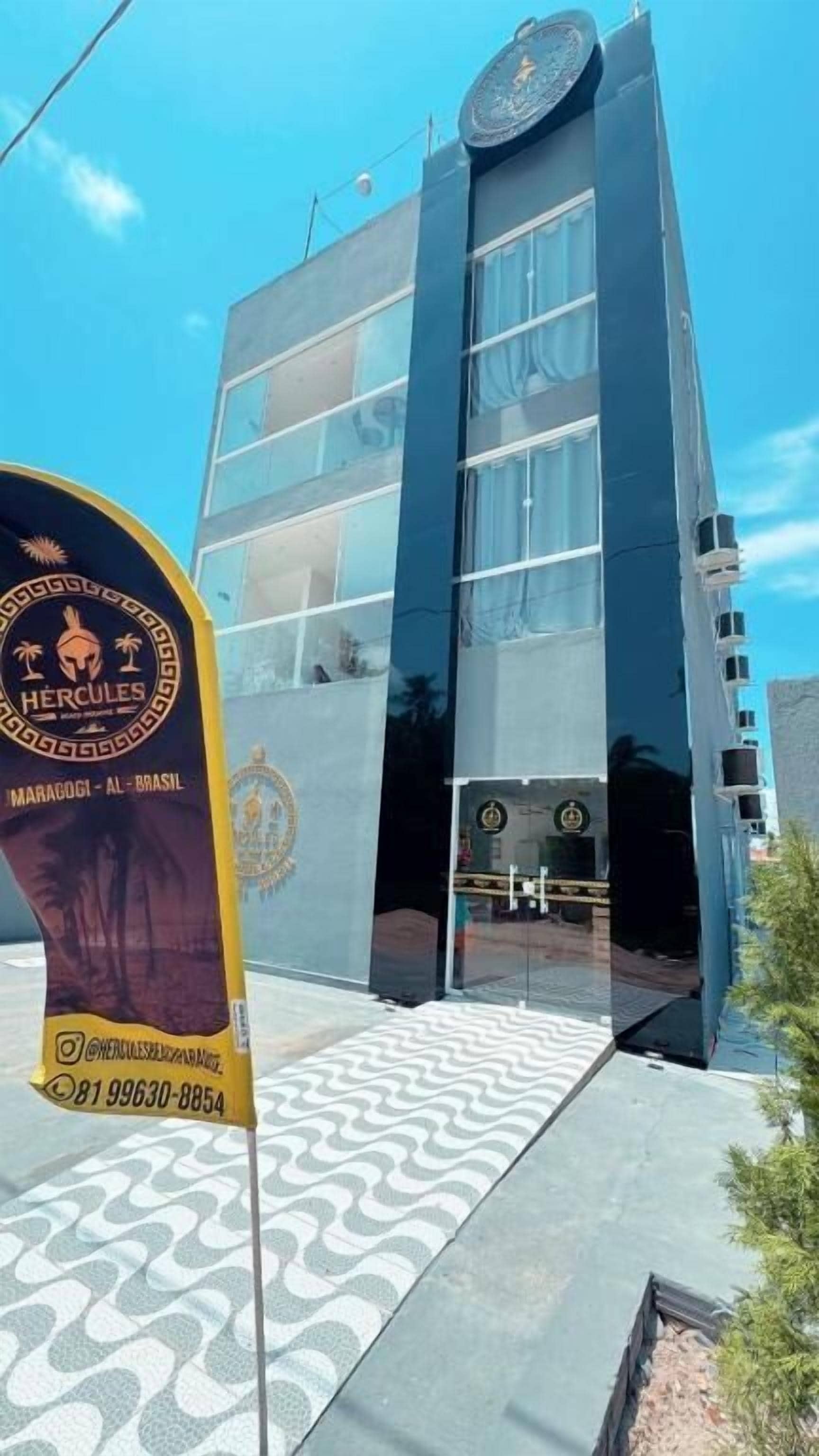 Un edificio moderno con fachada de vidrio, un logotipo con la palabra 'HERCULES' y un número de teléfono, y un sendero con diseño que conduce a la entrada.
