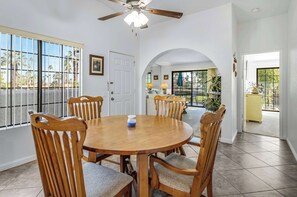 A dining area with a round wooden table and chairs, a ceiling fan, and a view through an arched doorway to a patio with plants.