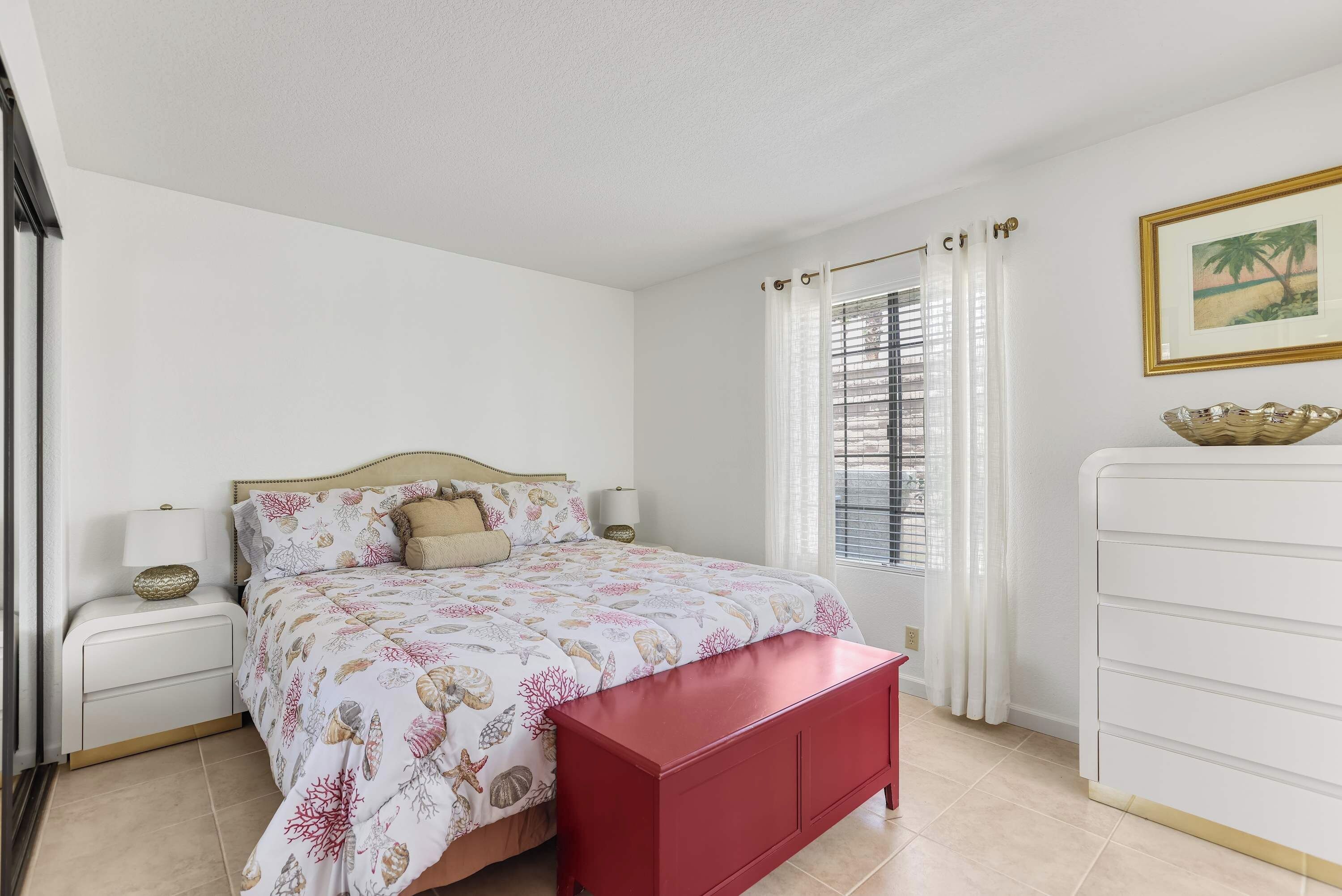 A bedroom with a bed, nightstands, a red bench, a dresser, and a window with curtains.