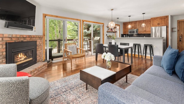 A modern living room with a fireplace, a sofa, a coffee table, and a dining area with chairs and a kitchen in the background. - Woodbridge 04A (Snowmass Village)