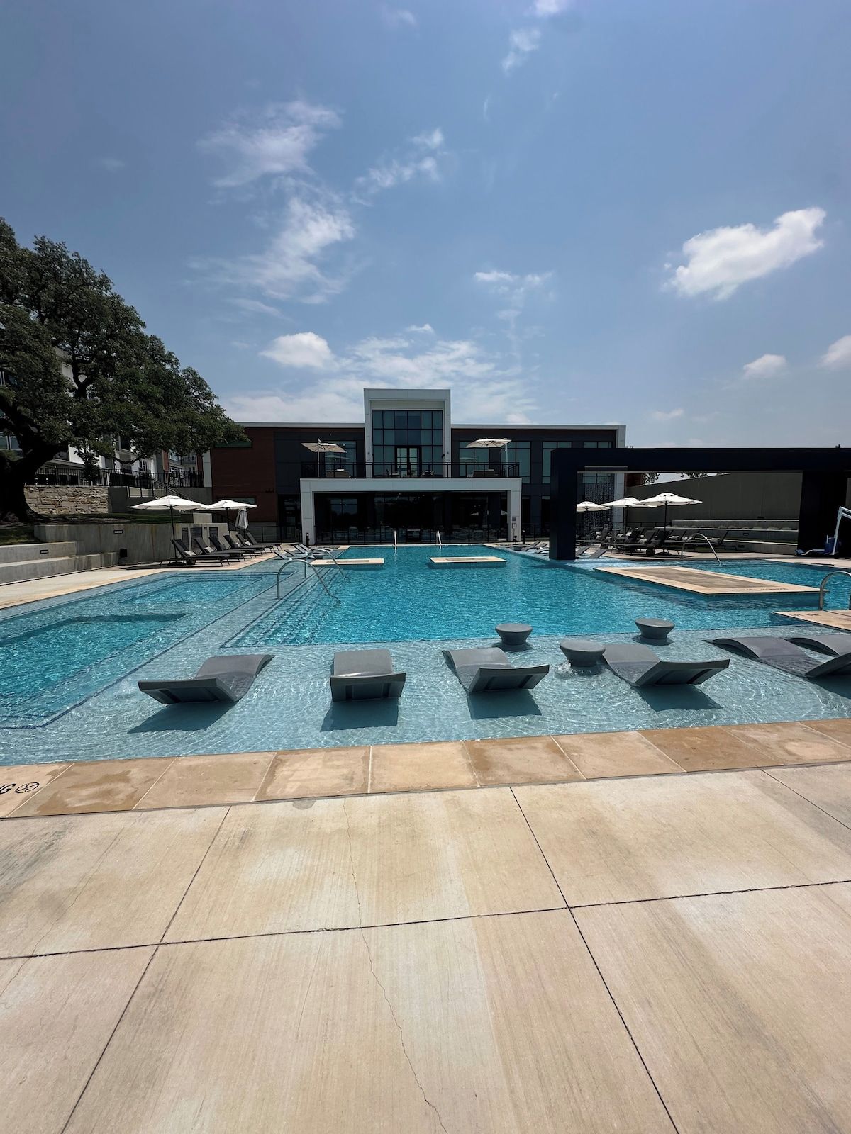 Piscina com espreguiçadeiras e guarda-sóis, cercada por um edifício com grandes janelas.