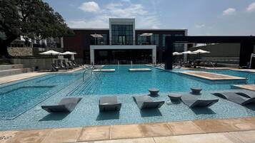 Piscina com espreguiçadeiras e guarda-sóis, cercada por um edifício com grandes janelas.