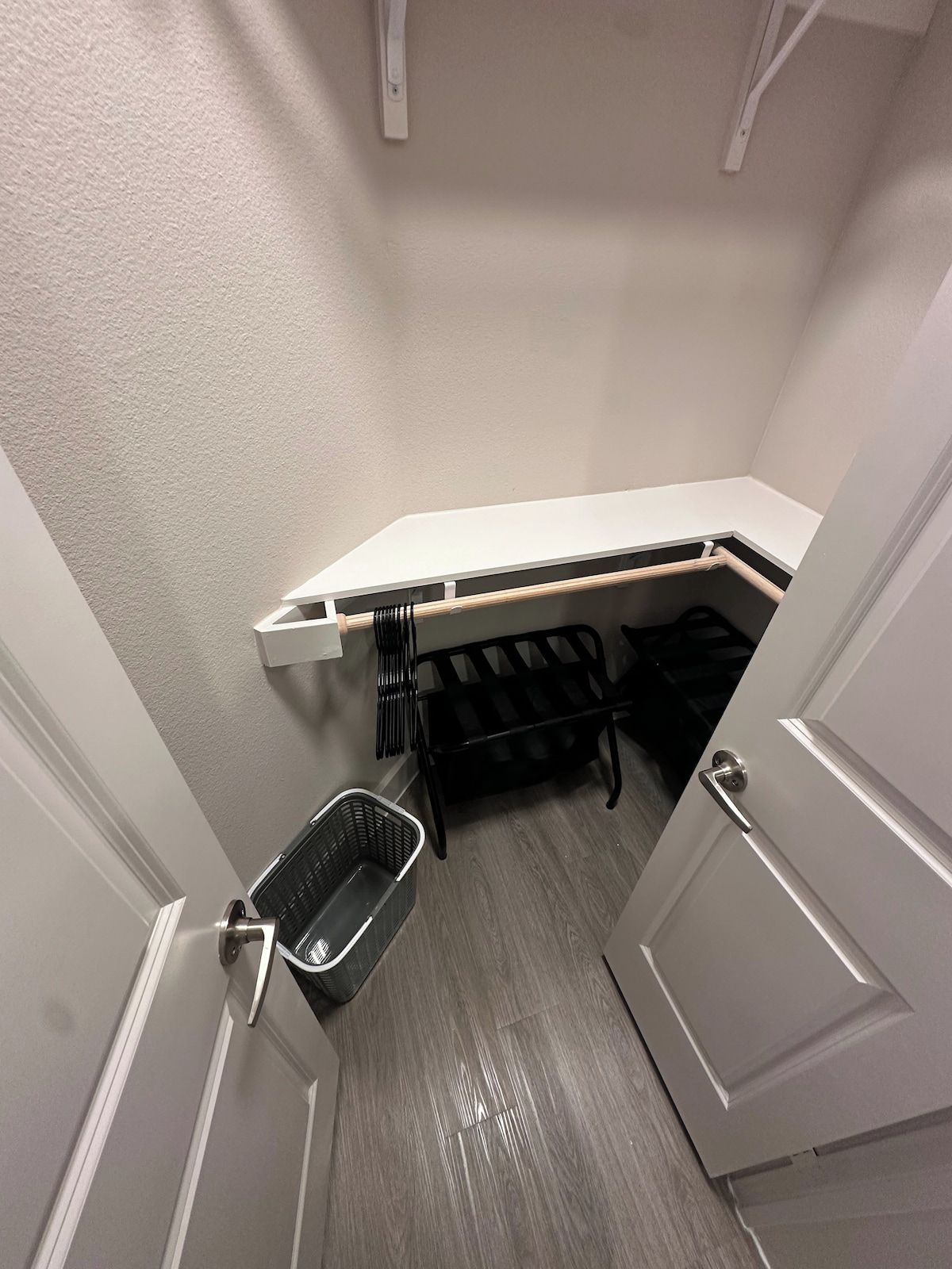 A compact room with a white door, a small trash bin, a black bench, and a white shelf unit.