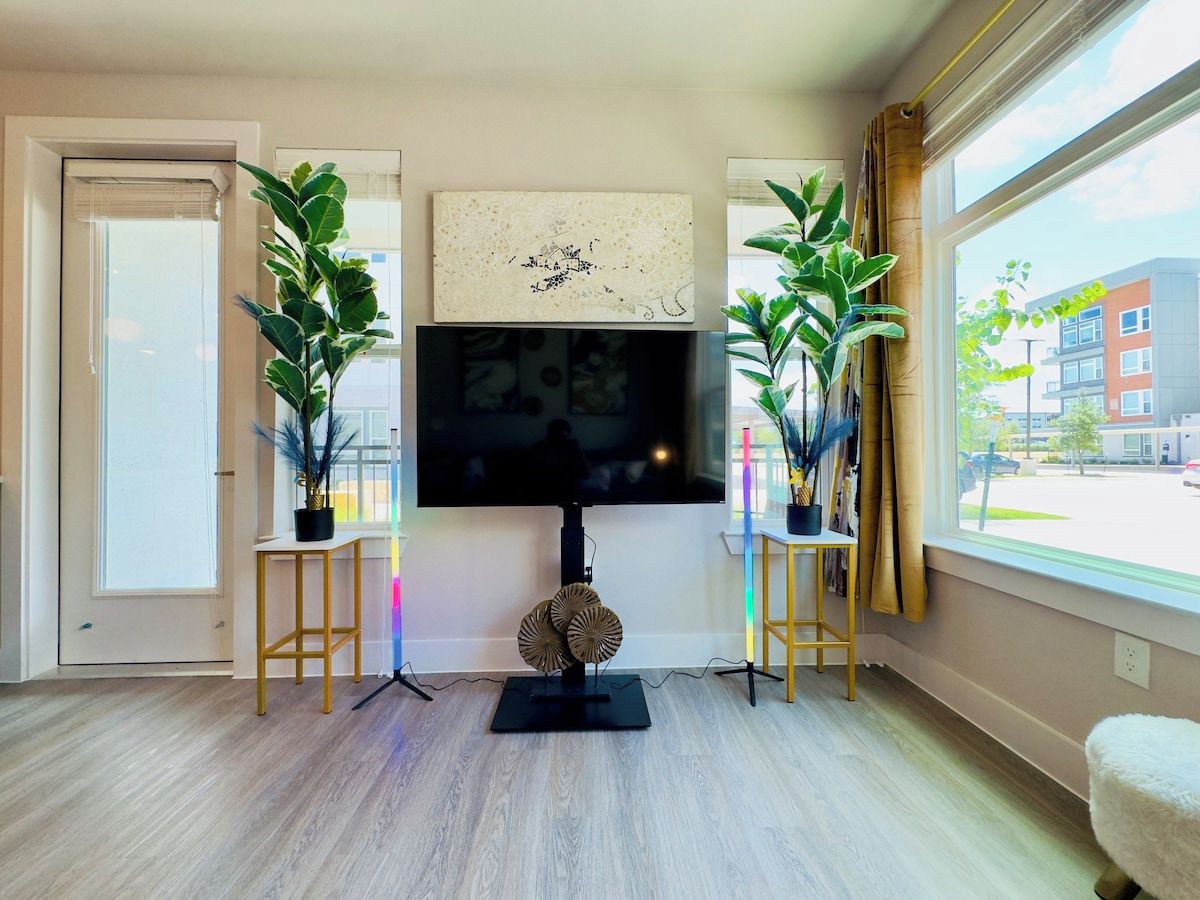 Un salon équipé d’un téléviseur à écran plat, de deux plantes en pot, d’un objet décoratif rond et d’une fenêtre donnant sur des immeubles.