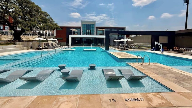 En pool med solstolar och parasoller, en modern byggnad i bakgrunden och en klar himmel.