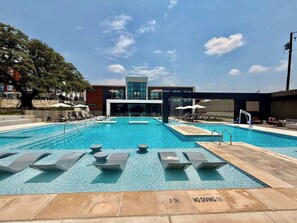 A swimming pool with lounge chairs and umbrellas, a modern building in the background, and a clear sky. - Longhorn-Themed 2BR | Near UT Austin & Q2 Stadium (Austin)