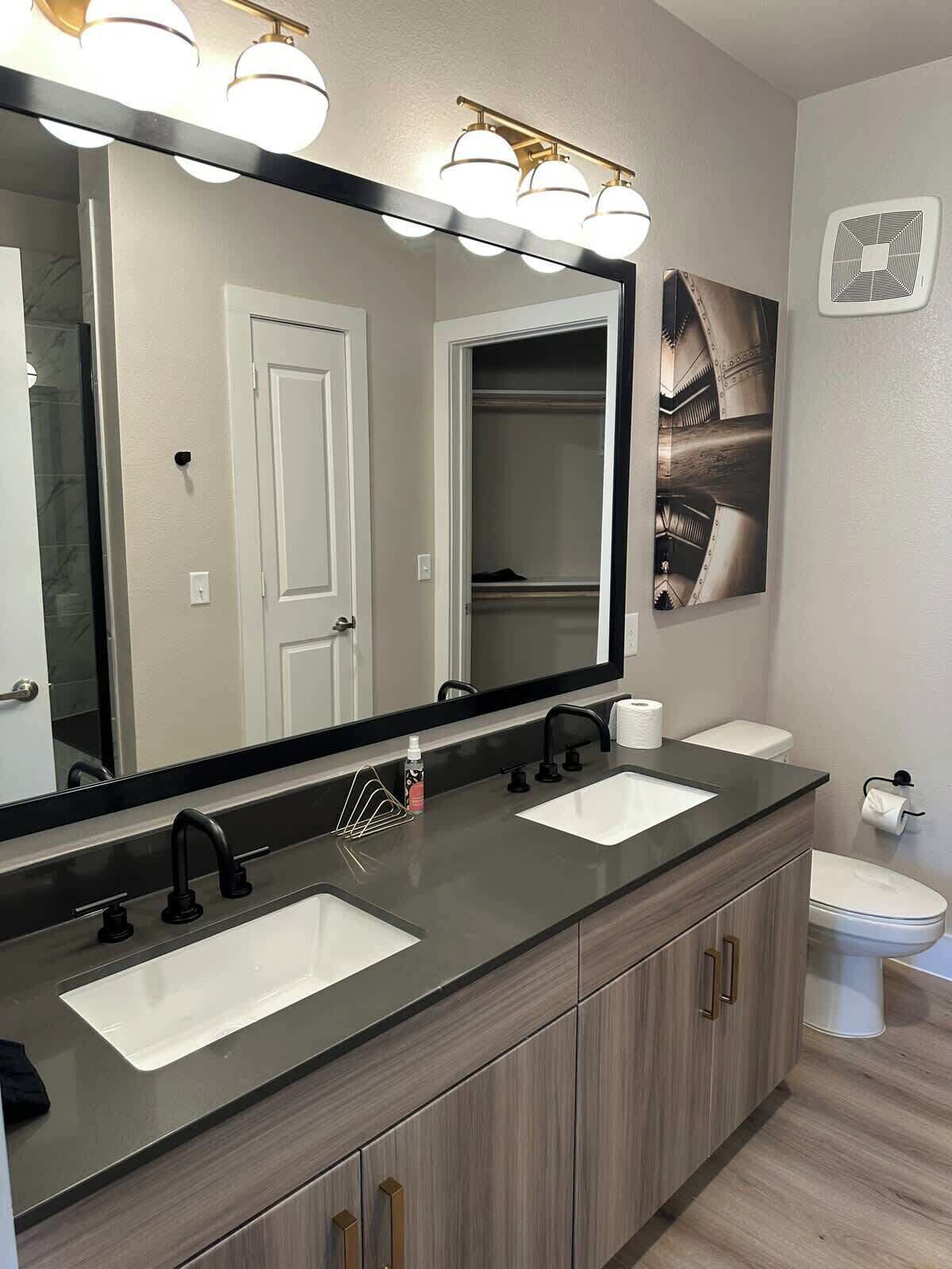 A modern bathroom with a large mirror, two sinks, a toilet, and a walk-in closet.