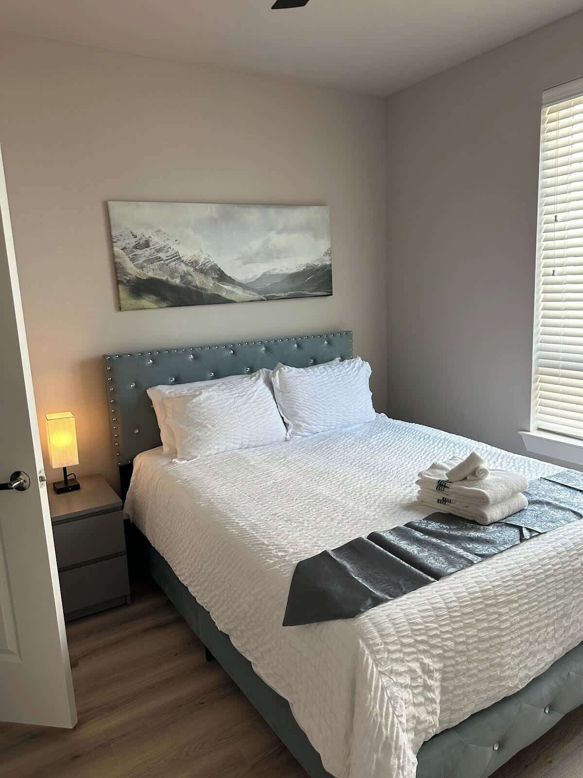 A bedroom with a bed, a nightstand, a lamp, a painting, and a window with blinds.