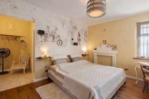 A bedroom with a large bed, a chair, a desk, a fireplace, and wall art.