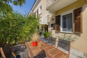 An outdoor dining area with a wooden table and chairs, surrounded by potted plants and a building with shutters.