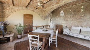 Una habitació rústica amb taula de fusta, cadires blanques, un sofà i sostre de rajola.