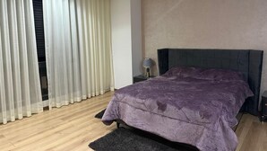 Un dormitori amb llit, una tauleta de nit, una làmpada i una finestra amb cortines translúcides.