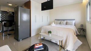 A modern bedroom with a large bed, a bedside table, a small bedside plant, and a flat-screen TV mounted on the wall.