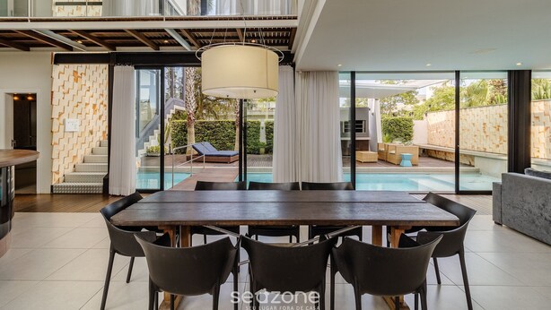 A modern dining area with a large table and chairs, a hanging light fixture, and a view of a pool and outdoor seating.