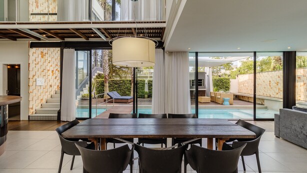 Un espace repas moderne avec une grande table et des chaises, un luminaire suspendu et une vue sur une piscine et un coin salon extérieur.