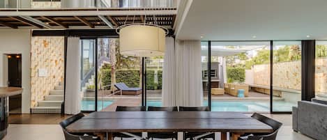 Un comedor moderno con una gran mesa y sillas, una lámpara colgante, y vistas a una piscina y zona de estar exterior.