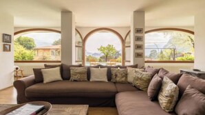 A living room with a large brown sofa, multiple cushions, and a view through arched windows to the outdoors.