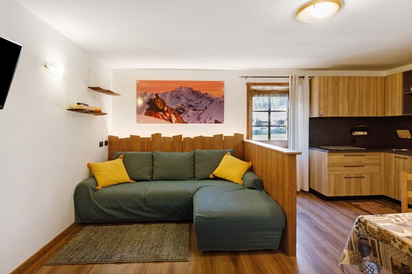 A room with a green sectional sofa, wooden flooring, a mounted flat-screen TV, a kitchen area with wooden cabinets, and a window with curtains.