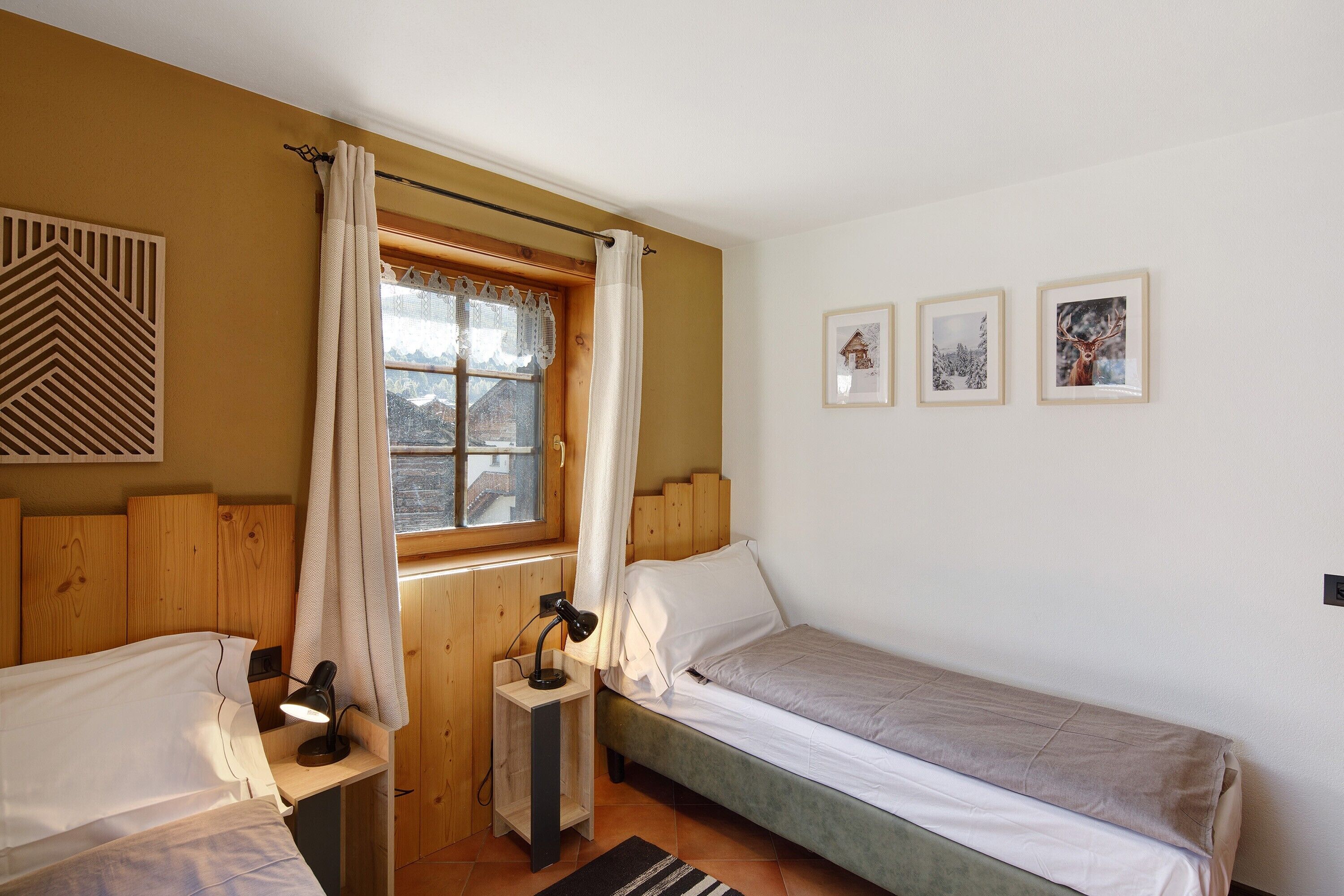 Een kleine, gezellige kamer met een enkel bed, een nachtkastje met een lamp, een raam met gordijnen en drie ingelijste foto’s aan de muur.