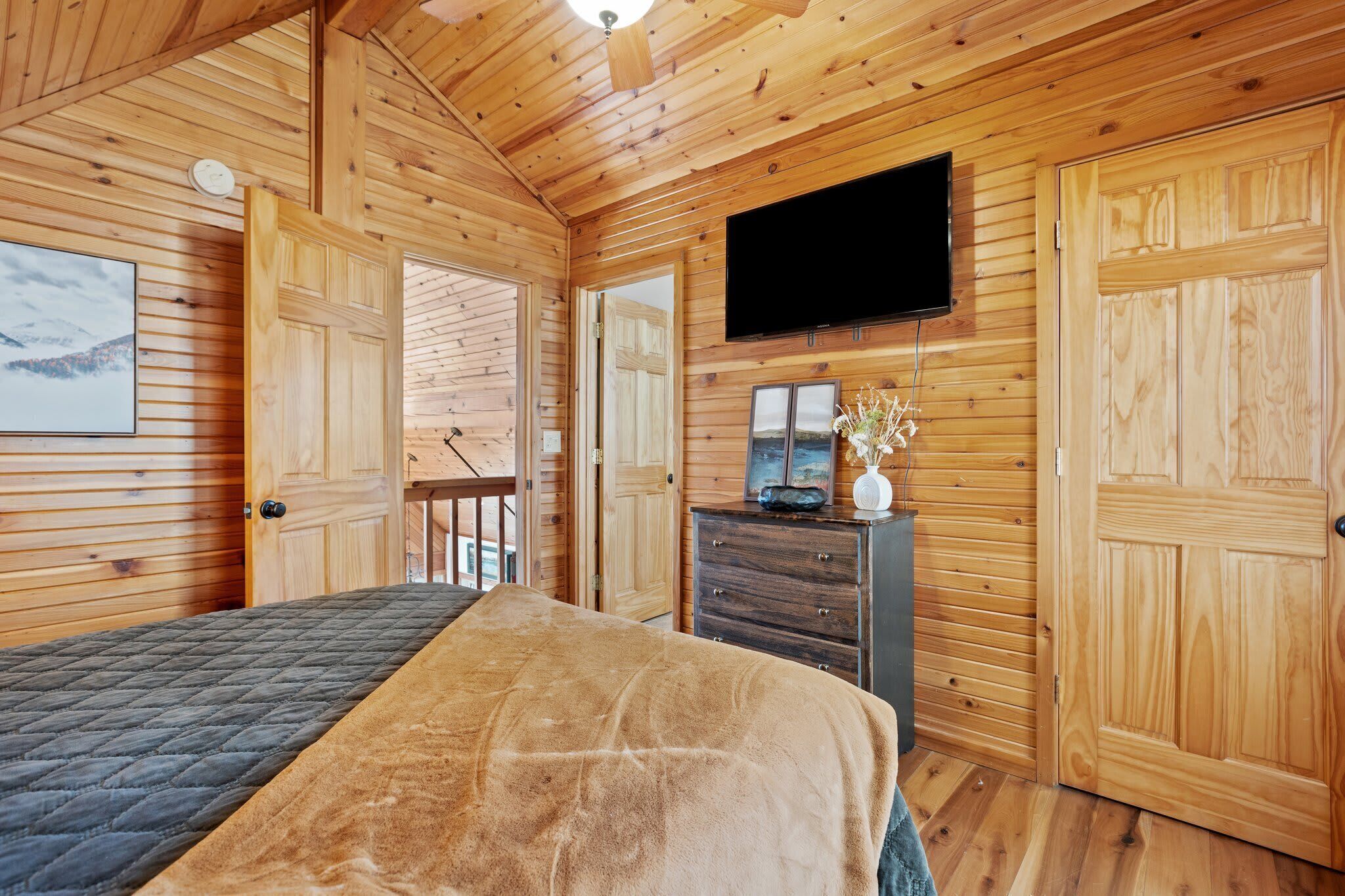 Habitación de cabaña de madera con cama, cómoda, televisor y puerta que da a un balcón.