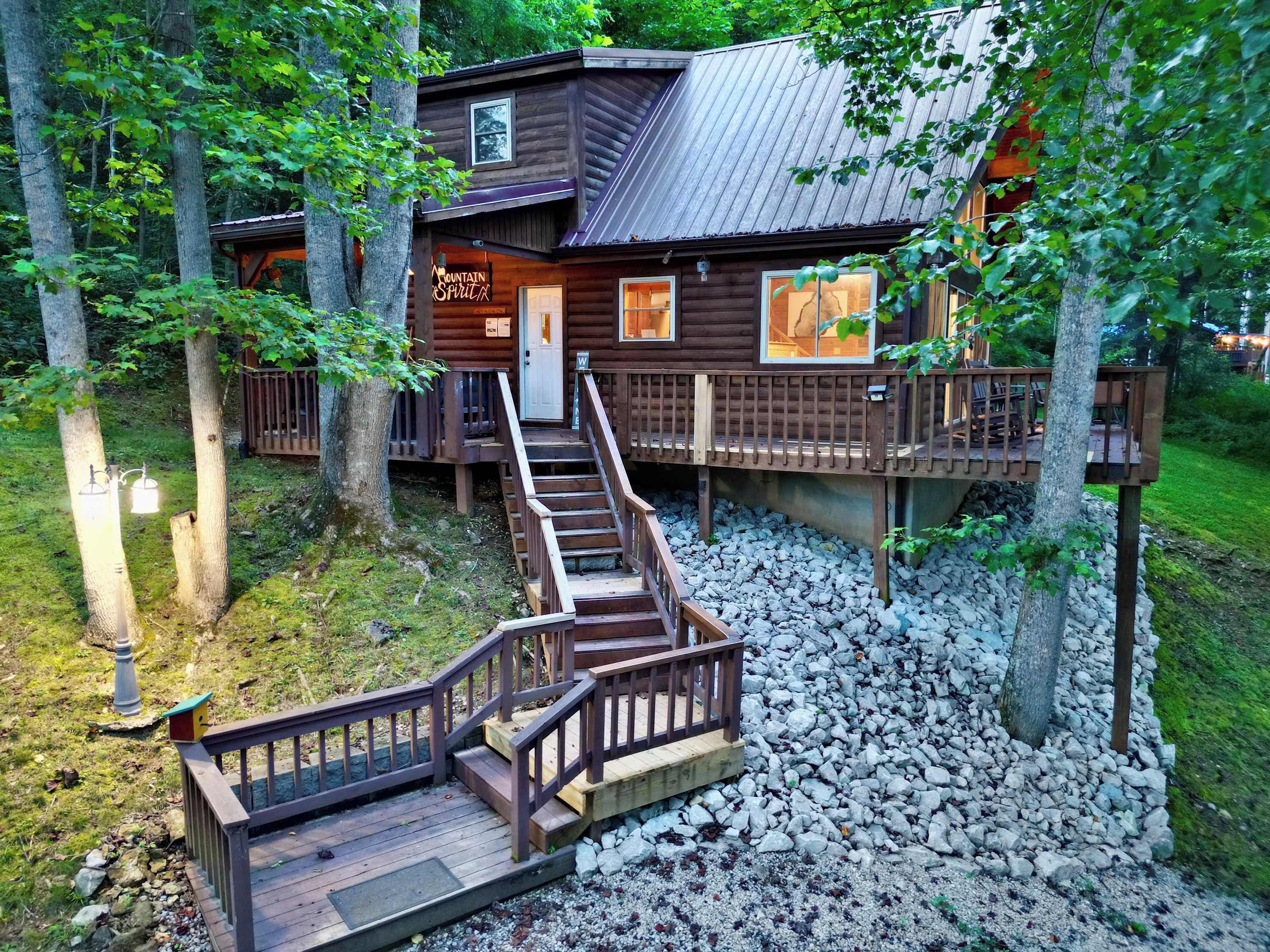 A wooden cabin with a staircase leading to a deck, surrounded by trees and gravel.