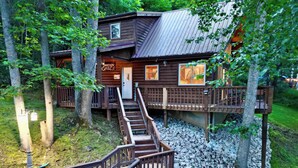Een houten chalet met een trap naar een terras, omgeven door bomen en grind.