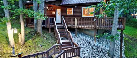 A wooden cabin with a staircase leading to a deck, surrounded by trees and gravel.