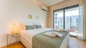 Ein modernes Hotelzimmer mit einem großen Bett, einem Nachttisch mit Lampe, einem roten Sessel am Fenster und einem Balkon mit Blick auf Gebäude.