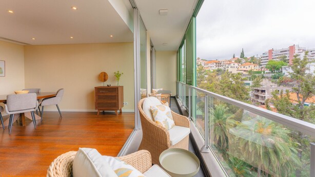 A balcony with wicker furniture, a dining area, and a view of a cityscape. - FLH Funchal City View at Quinta da Nogueira Reside (Funchal)