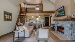 A spacious living area with a high ceiling, a stone fireplace, a kitchen with a central island, and a staircase leading to an upper level. - Cottage on the 10th Tee Beautiful Stay for 7 w/ Hot Tub & Fire Pit (Eureka)