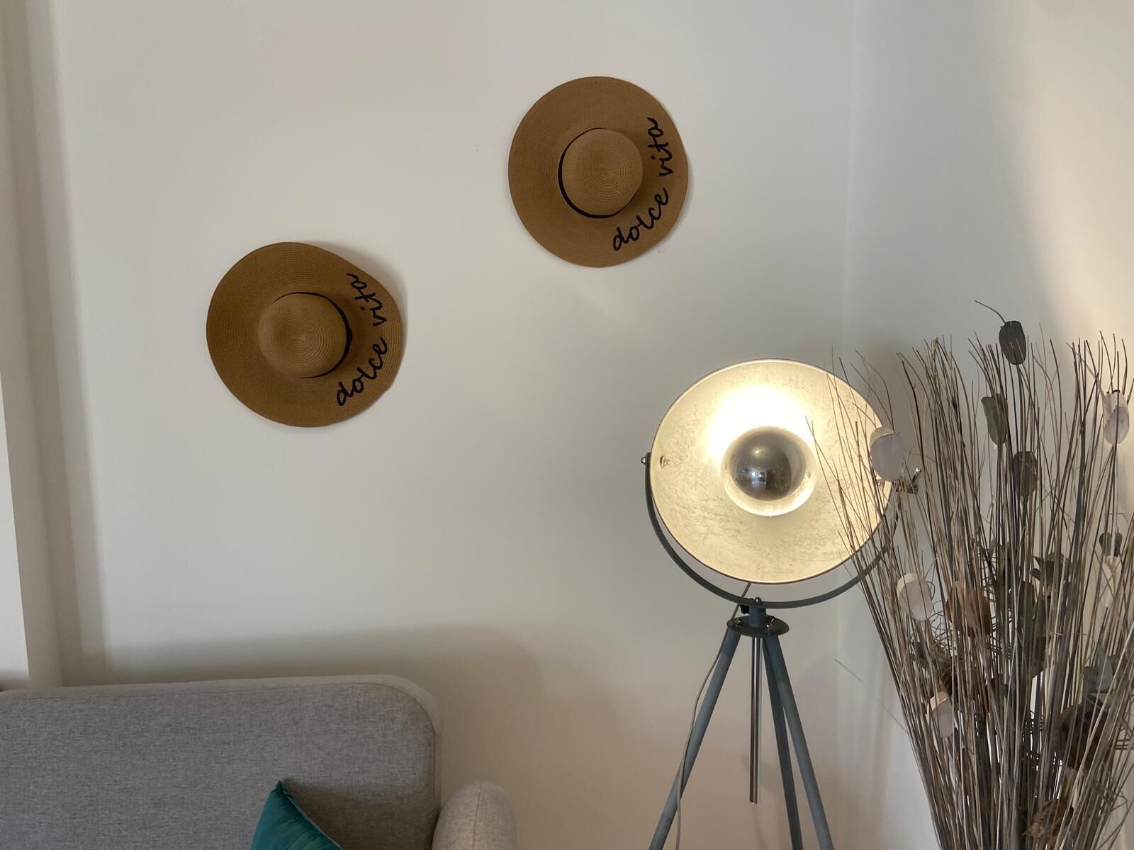 Een kamer met een grijze bank, een vloerlamp en drie hoeden aan de muur, met de tekst 'dolce' erop.