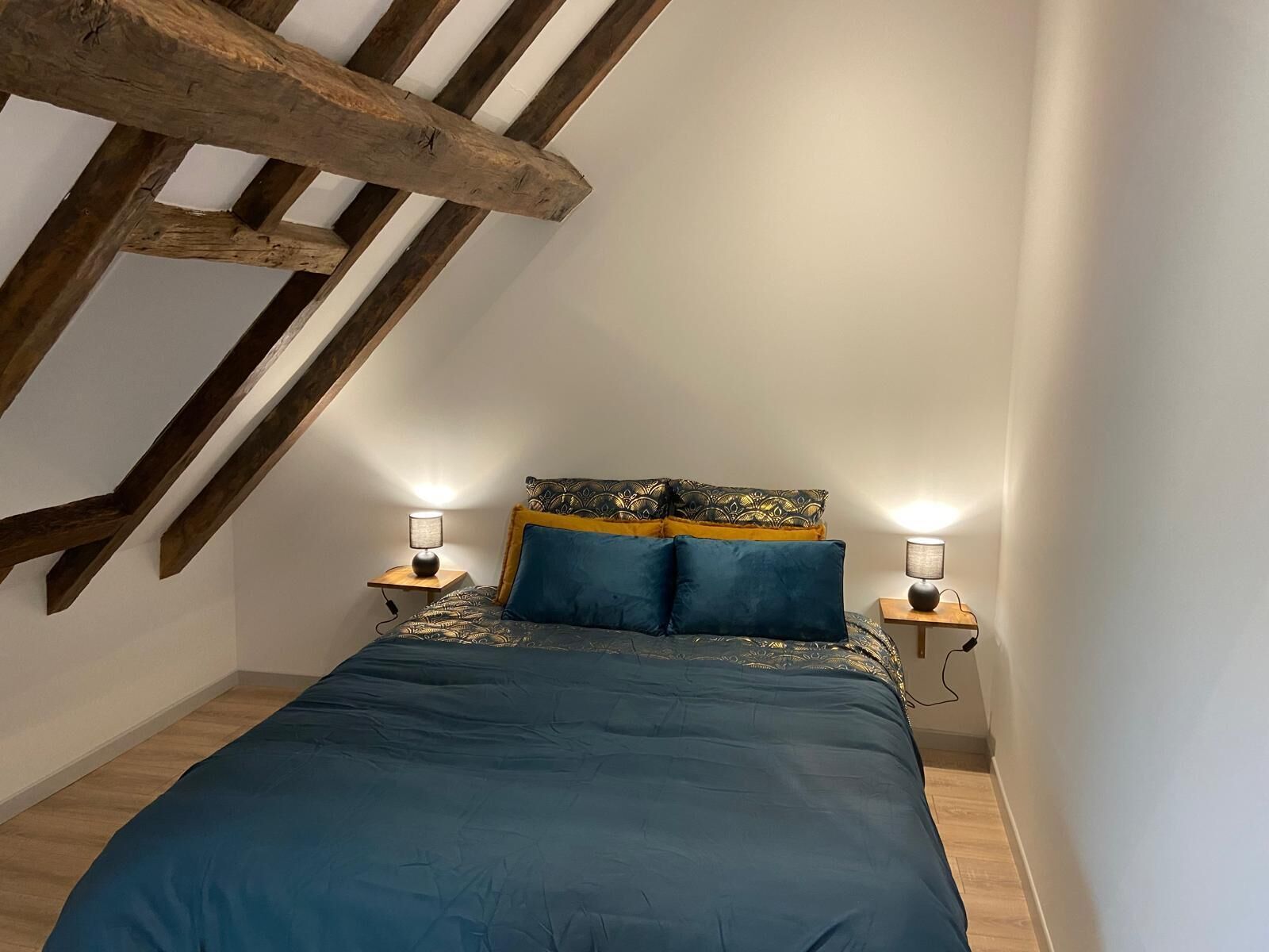 Ein Schlafzimmer mit schrägen Wänden, Holzbalken, einem Bett mit blauer Bettwäsche, zwei Nachttischen mit Lampen und gemusterten Kissen.