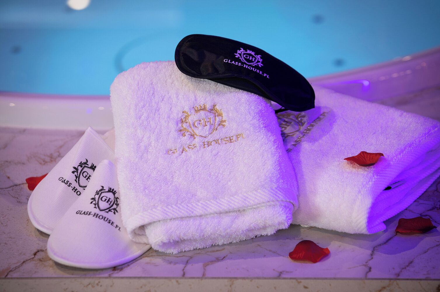 Un ensemble de serviettes et une casquette portant le logo « Glass-House.PL » sont posés sur une surface en marbre, près d’une piscine.