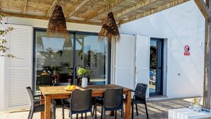 An outdoor dining area with a wooden table and chairs, a hanging basket, and a glass door leading to a pool area.