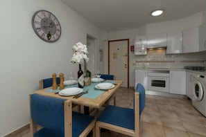 A dining area with a wooden table set for four, blue chairs, a wall clock, and a kitchen with white cabinets and appliances.